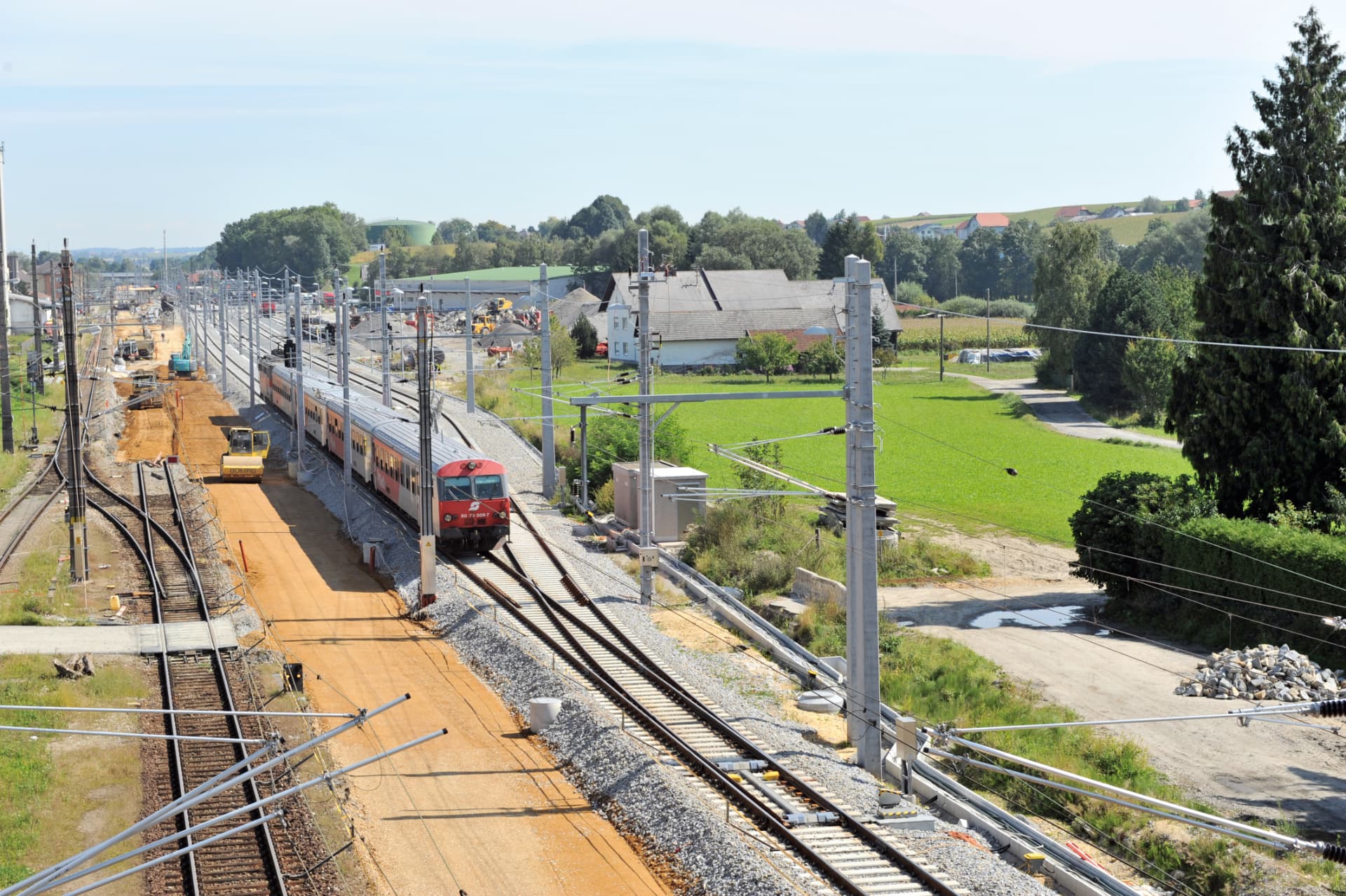 Blick auf mehrere Gleise mit Oberleitungsmasten, Auslegern und Fahrdraht; ein Triebwagen steht neben einer Gleisbaustelle